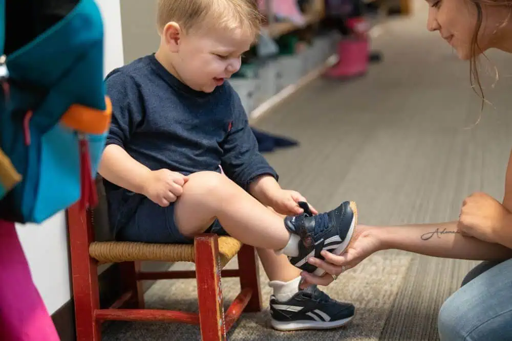 Toddler putting on shoes with a kind Christian Montessori caregiver helping