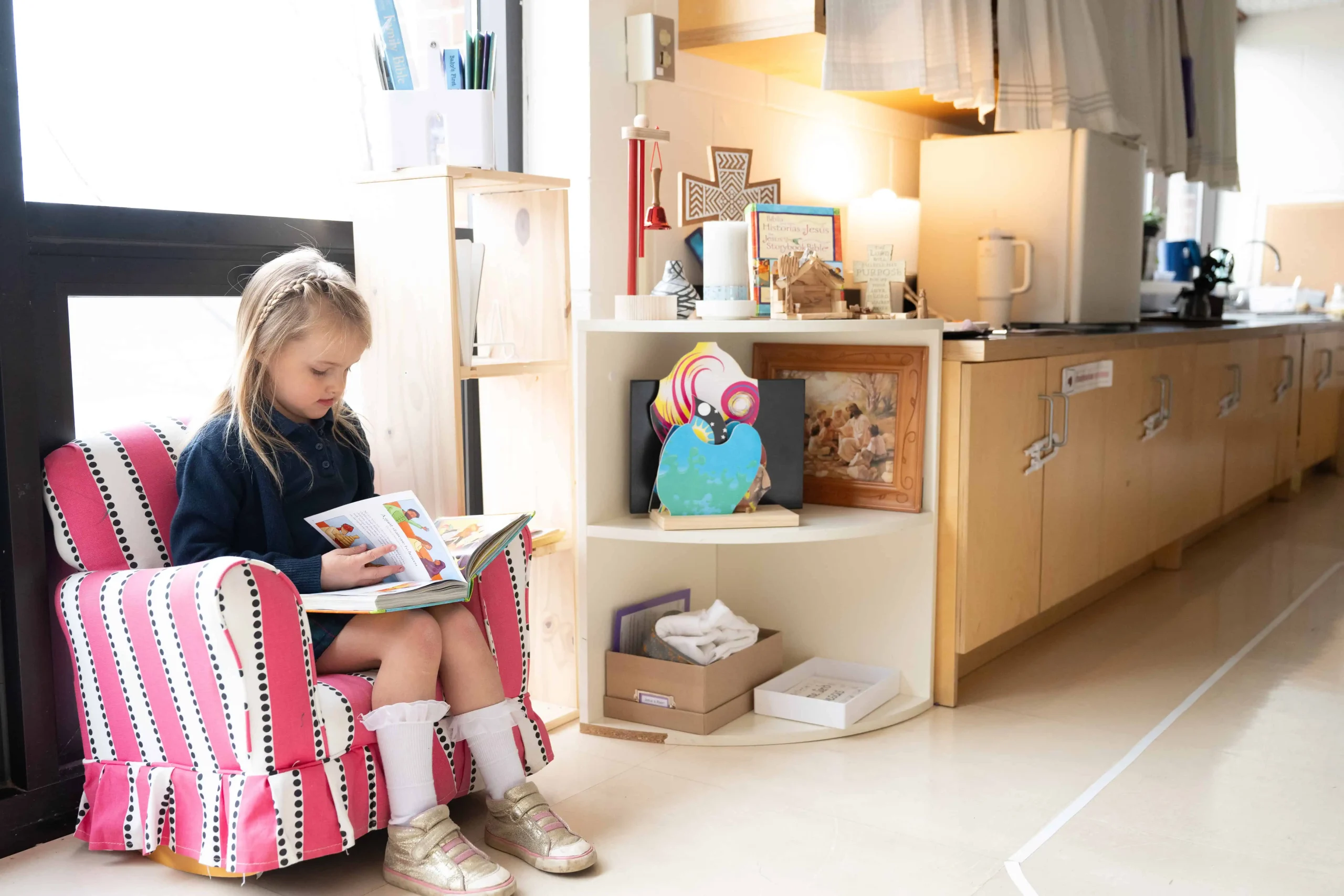 Kindergarten student reading in child sized chair at Hand In Hand Christian Montessori